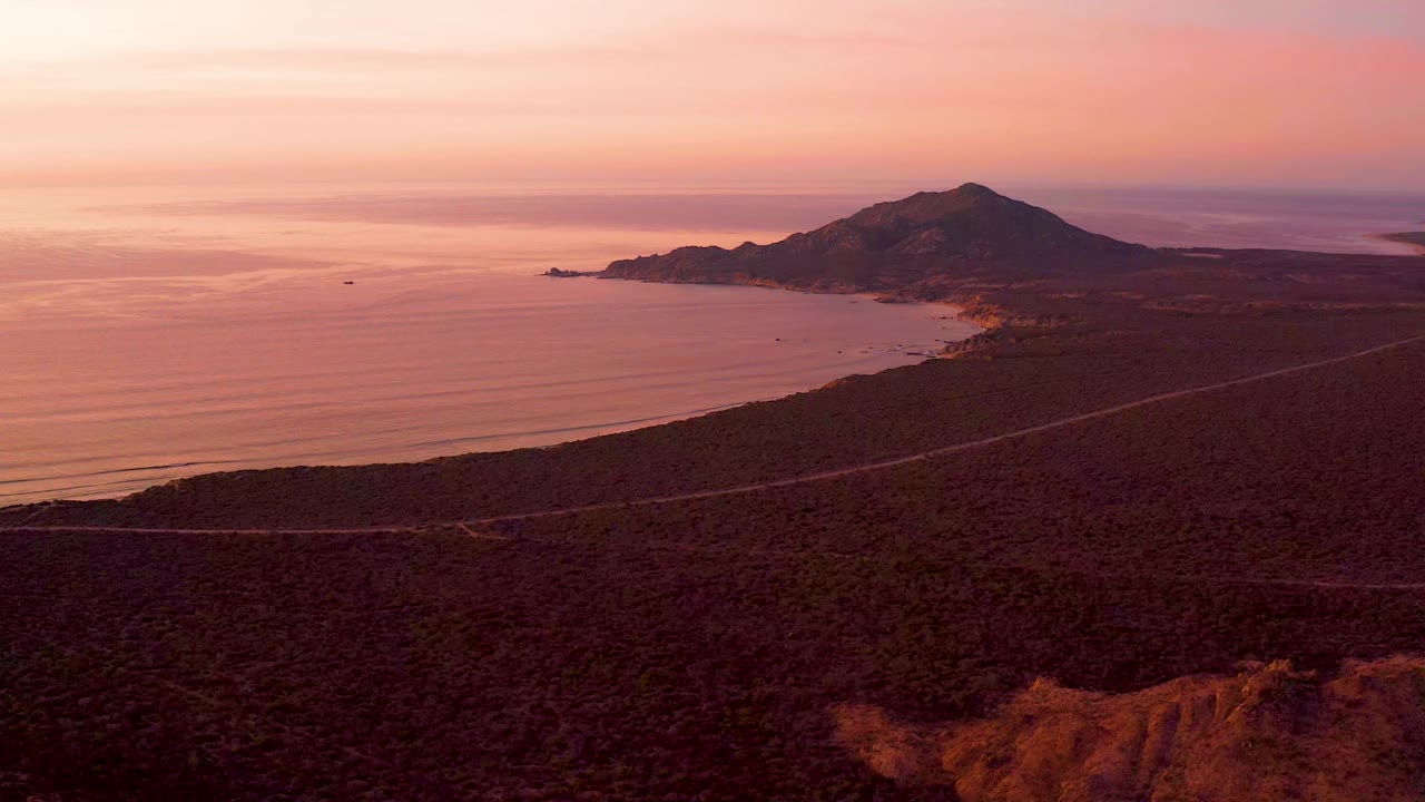 Cabo Pulmo aerial view one second in, with the mountain centered beyond the coastline, soft ocean bands to the left, a road across the dark foreground, and no visible people.