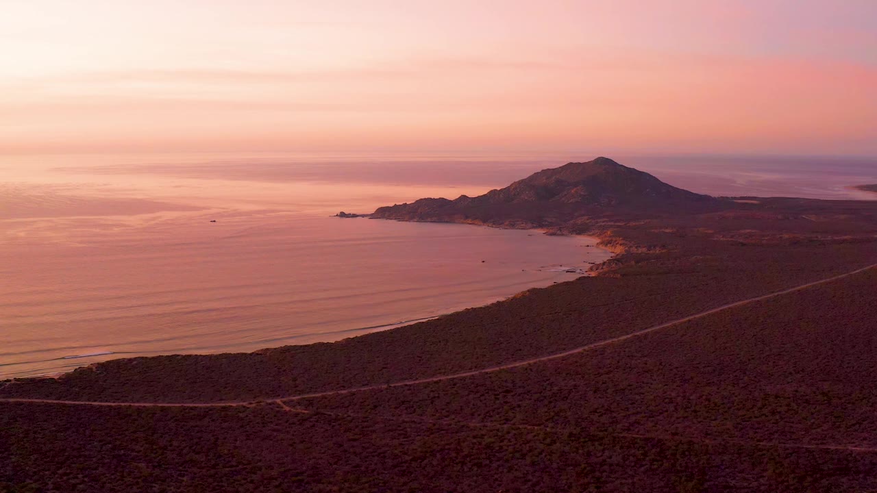 Elevated Cabo Pulmo view with the road crossing dry terrain below, calm water along the shore, the mountain centered in the background, and no visible people.