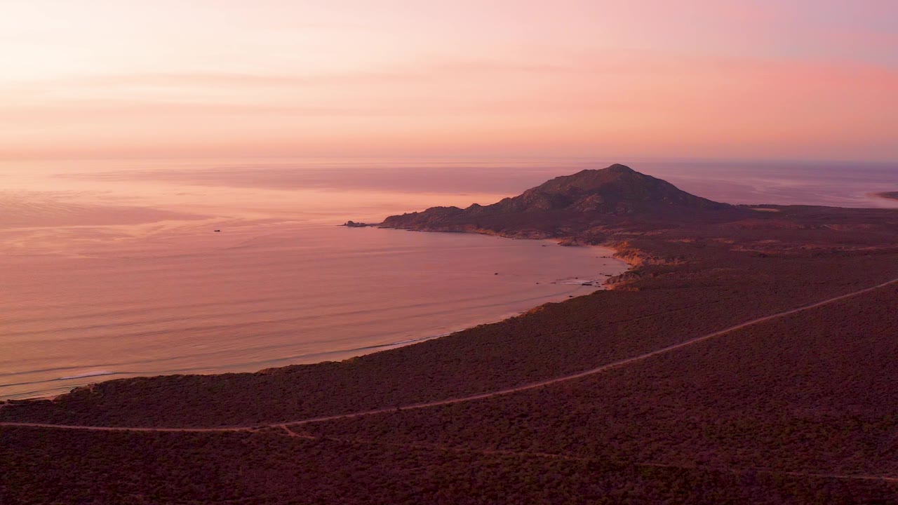 Wide Cabo Pulmo coastal view with the ocean filling the left side, a road crossing the lower dry slope, mountain ridges in the distance, and no visible people.