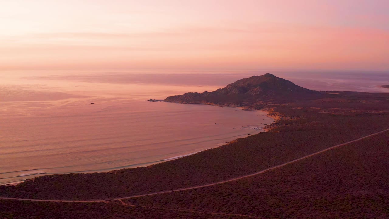 Cabo Pulmo coastline under pink dusk light, showing the ocean to the left, low beach at the bottom edge, dark mountain mass to the right, a diagonal road across scrubland, and no visible people.