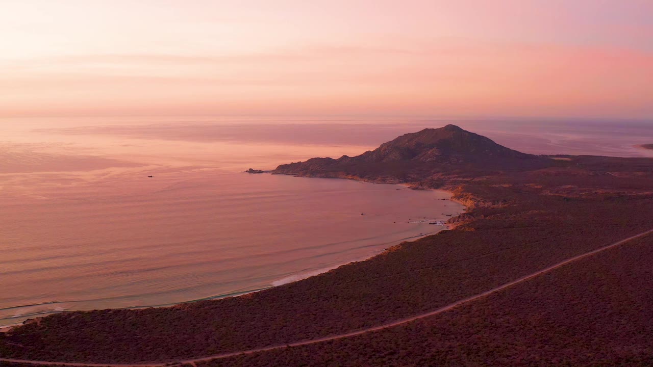 Cabo Pulmo coastline seen from above with the mountain slightly right of center, smooth sunset-colored water to the left, a diagonal road along the shore, and no visible people.