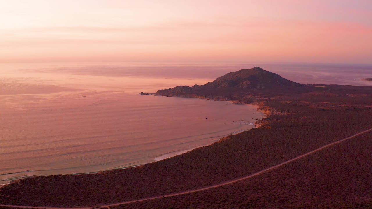 Cabo Pulmo sunset view with more open water across the left half of frame, a dark mountain ridge along the shore, the road visible in the lower right, and no visible people.