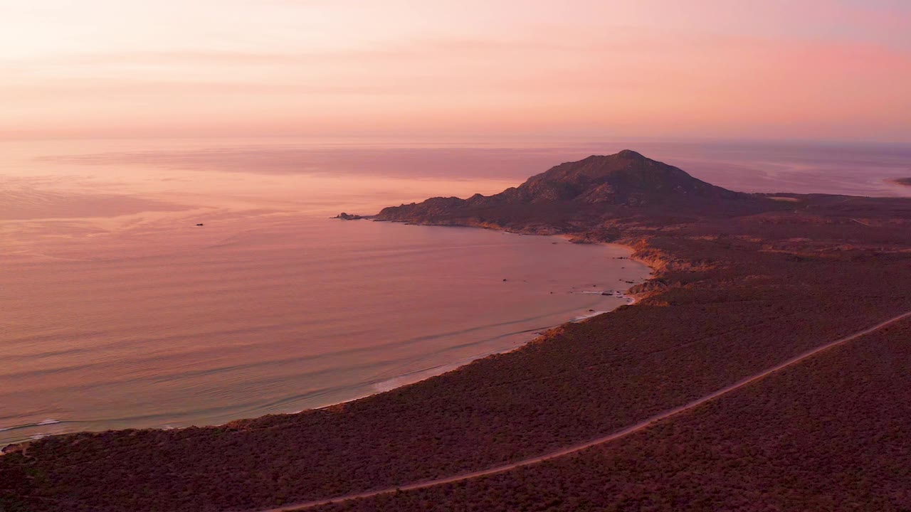 Cabo Pulmo coastline at 0:01 with the ocean reflecting pink light, a diagonal road across the dark foreground, the shoreline curving toward a mountain, and no visible people.