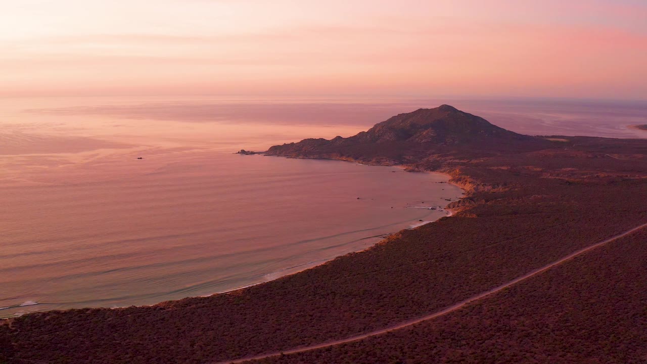 Cabo Pulmo bay at 0:03 with soft orange light on the water, a dark foreground hillside crossed by a road, the coastline bending toward the mountain, and no visible people.