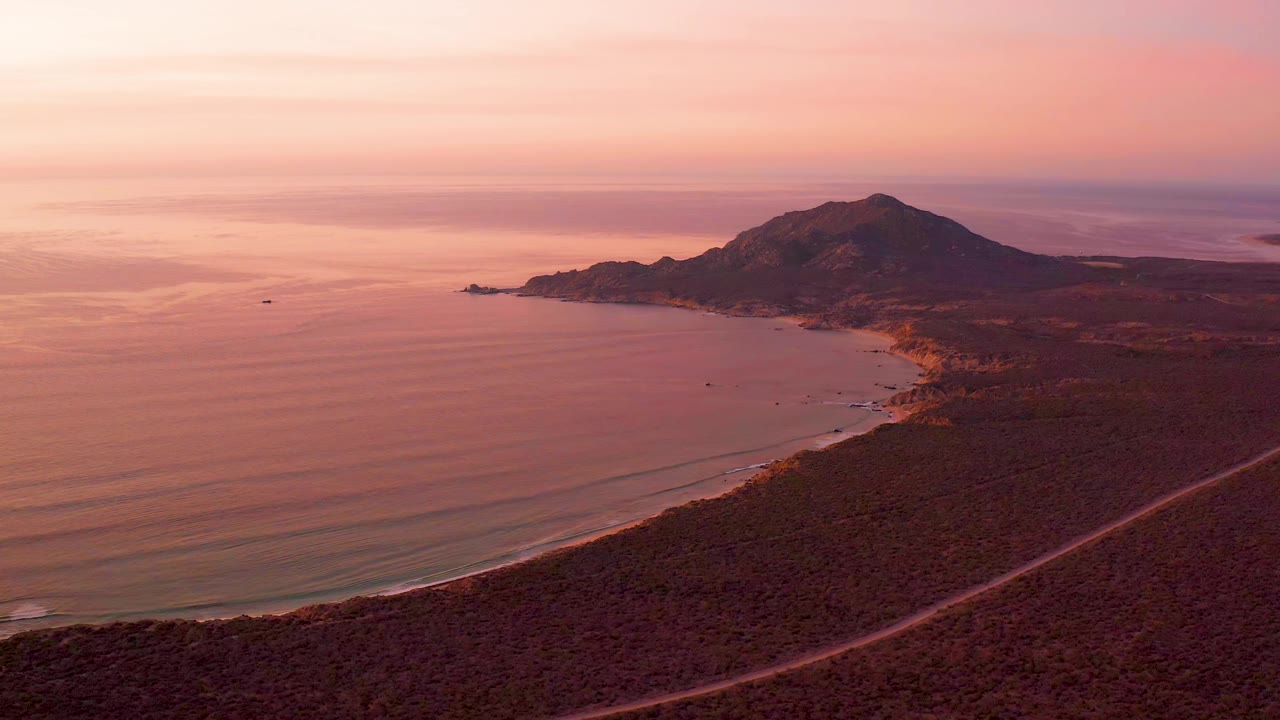 Cabo Pulmo shoreline seen from above at sunset, with the hill slightly right of center, faint parallel waves, the coastal road below, a small boat on the water, and no visible people.