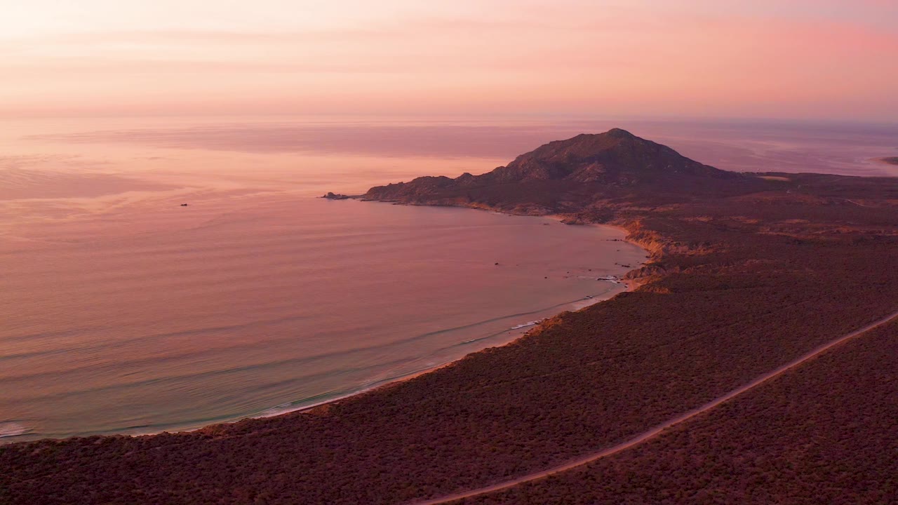 A warm sunset view over Cabo Pulmo shows the ocean on the left, the curving beach and road below, the hill rising near the shore, a small boat in the bay, and no visible people.