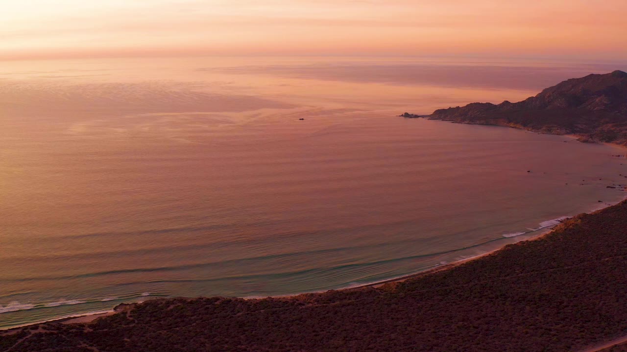 Aerial sunset view of ocean, beach, hillside, and distant coastal mountains.