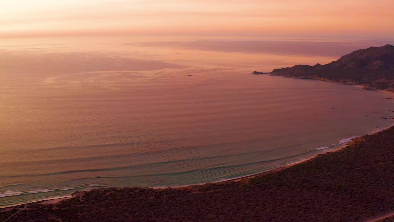 Cabo Pulmo coastline at 0:01 with the curved beach lower in frame, calm sea filling most of the view, layered sunset color in the sky, right-side mountains, and no visible people.