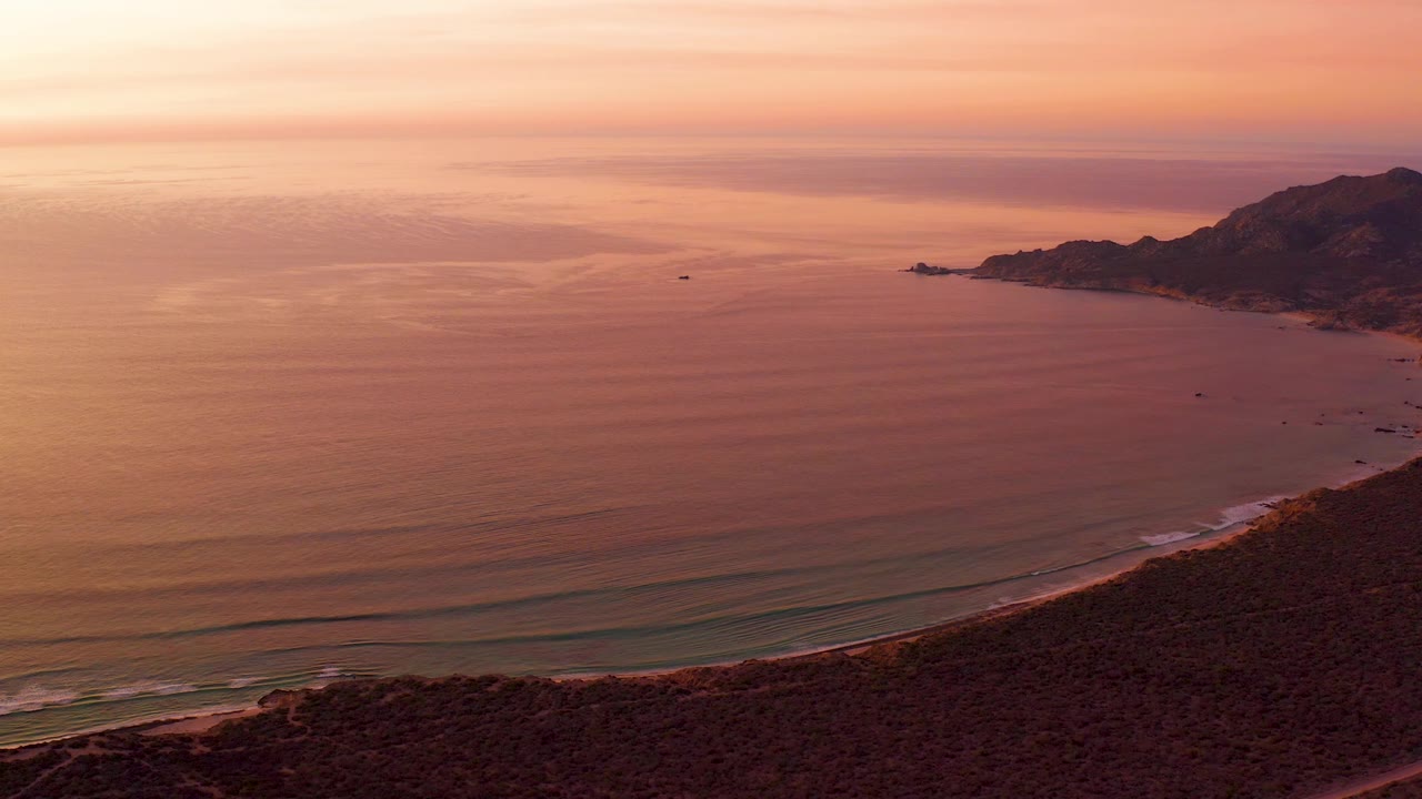 Cabo Pulmo aerial view at 0:03 with open ocean across the left and center, the beach tracing the lower edge, a mountain headland on the right, soft orange-pink sky, and no visible people.