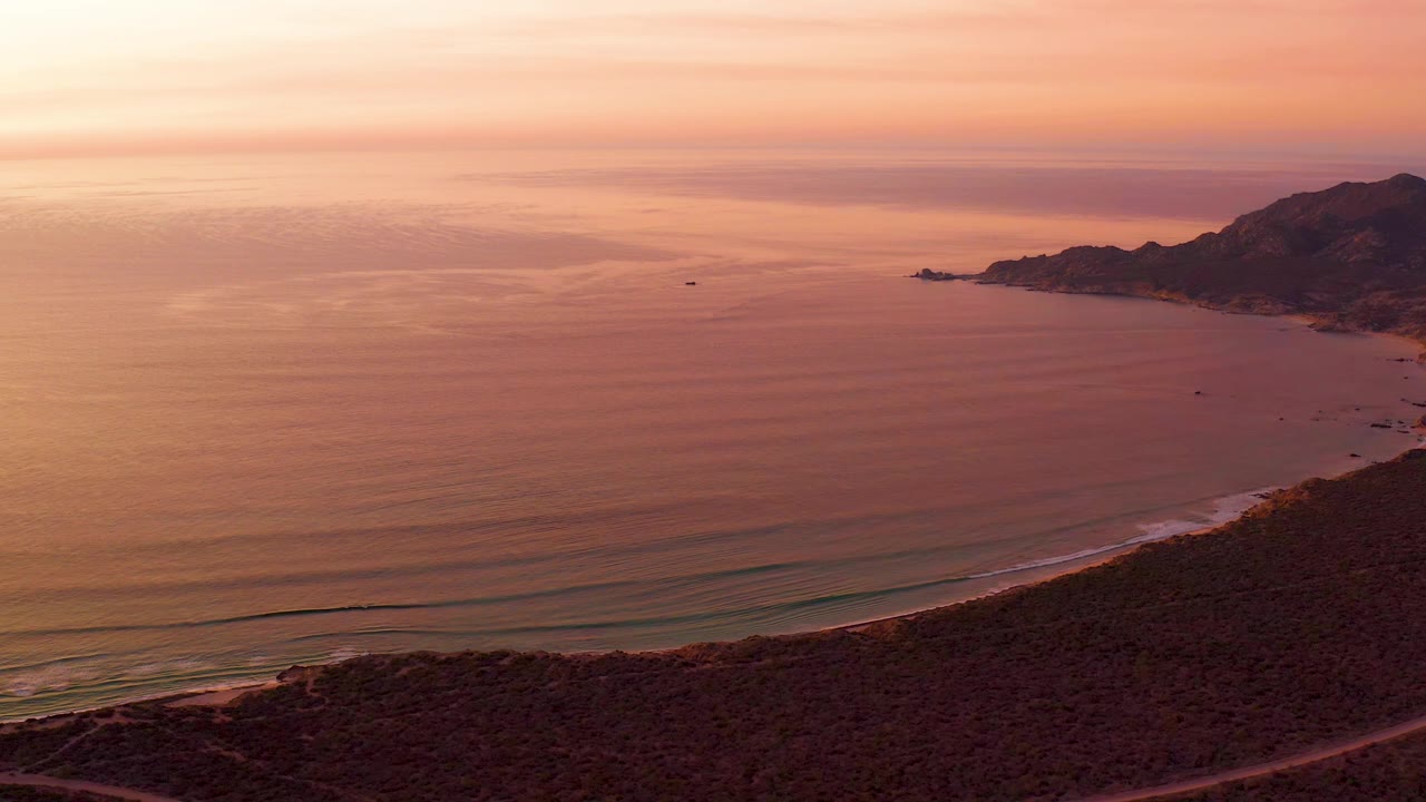 Wide Cabo Pulmo coastline frame with the ocean spread across the center, the road crossing the lower scrubland, mountains on the right horizon, warm dusk sky, and no visible people.