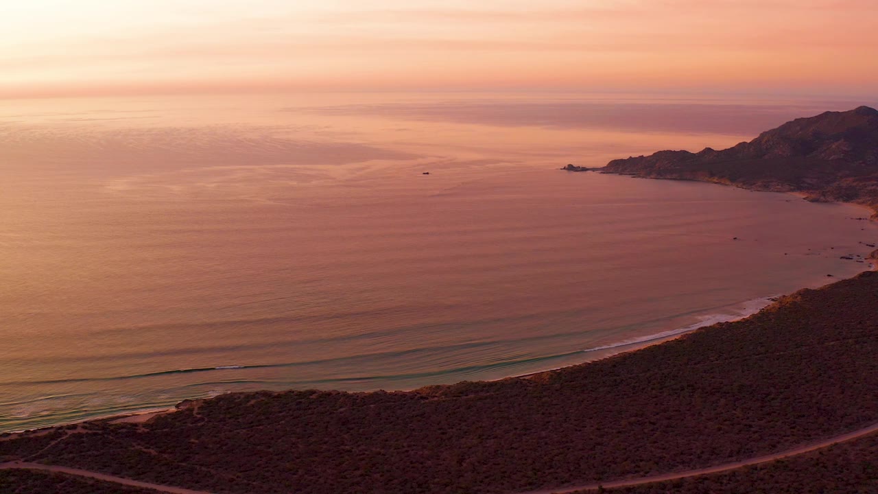 High view of Cabo Pulmo coast with the ocean taking most of the frame, a small road and scrubland along the lower edge, a beach with thin wave bands, distant mountains, and no visible people.