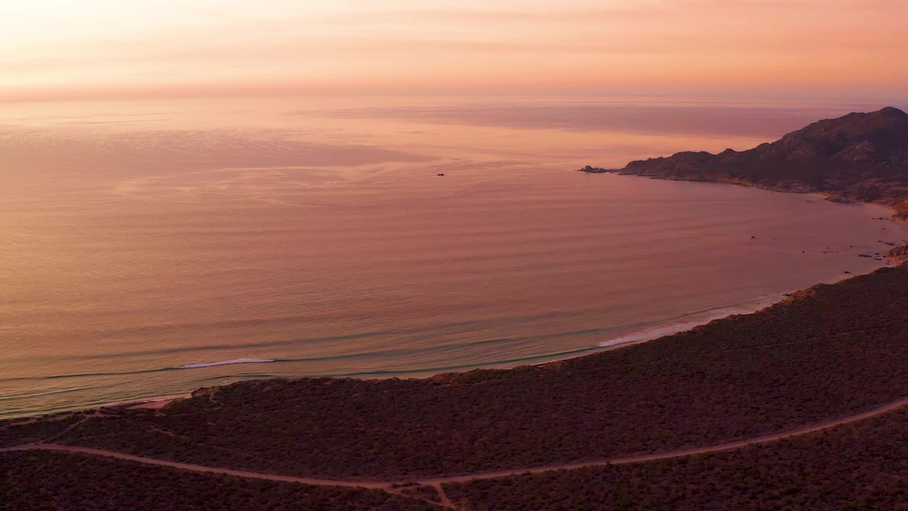 Wide aerial view in Cabo Pulmo showing orange-pink ocean water, parallel surf bands along the beach, a curved road through foreground scrub, right-side hills, and no visible people.