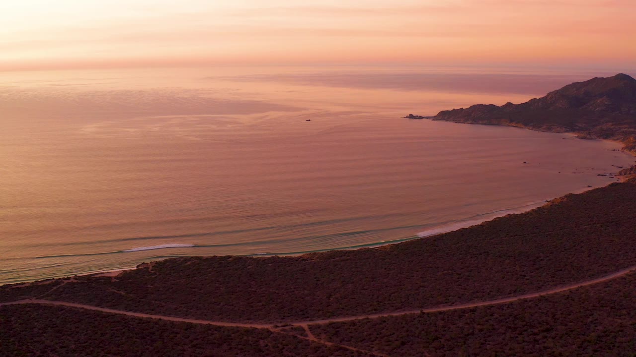 Aerial Cabo Pulmo view with a low foreground road, dark scrubland, a narrow beach line, soft wave sets, warm orange water, hills to the right, and no visible people.
