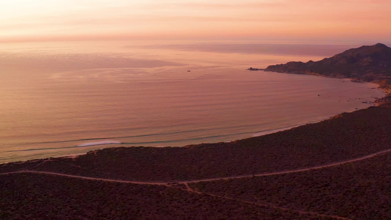 Coastal road crosses a scrub hillside below orange sunset water.