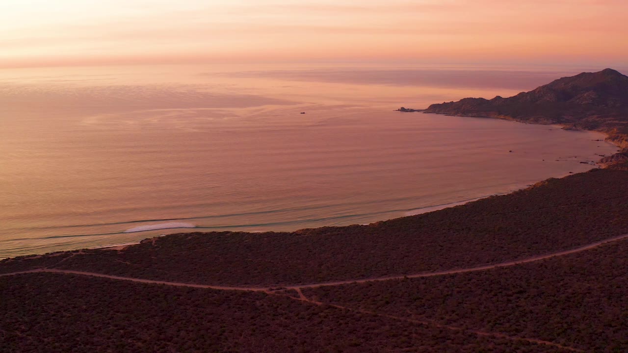 Cabo Pulmo aerial view with the foreground hillside darkened by dusk, a winding road along the slope, calm sea stretching to the horizon, right-side coastal mountains, and no visible people.