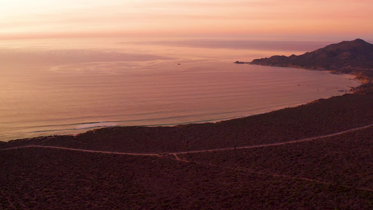 Aerial frame over Cabo Pulmo, Baja California Sur showing a winding road across dark hillside scrub, calm ocean beyond, distant hills to the right, warm sunset sky, and no visible people.