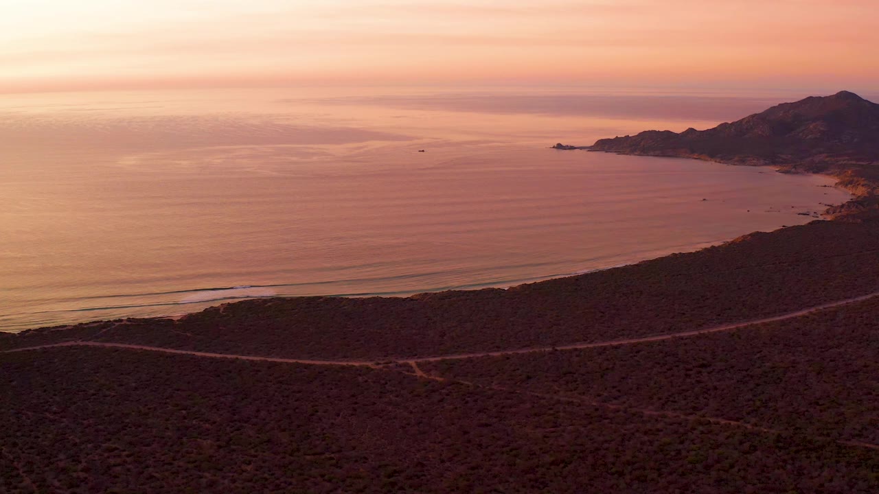 Elevated Cabo Pulmo coastal view with the road tracing the lower hillside, broad calm water in the center, small offshore specks, right-side hills, pink-orange sky, and no visible people.