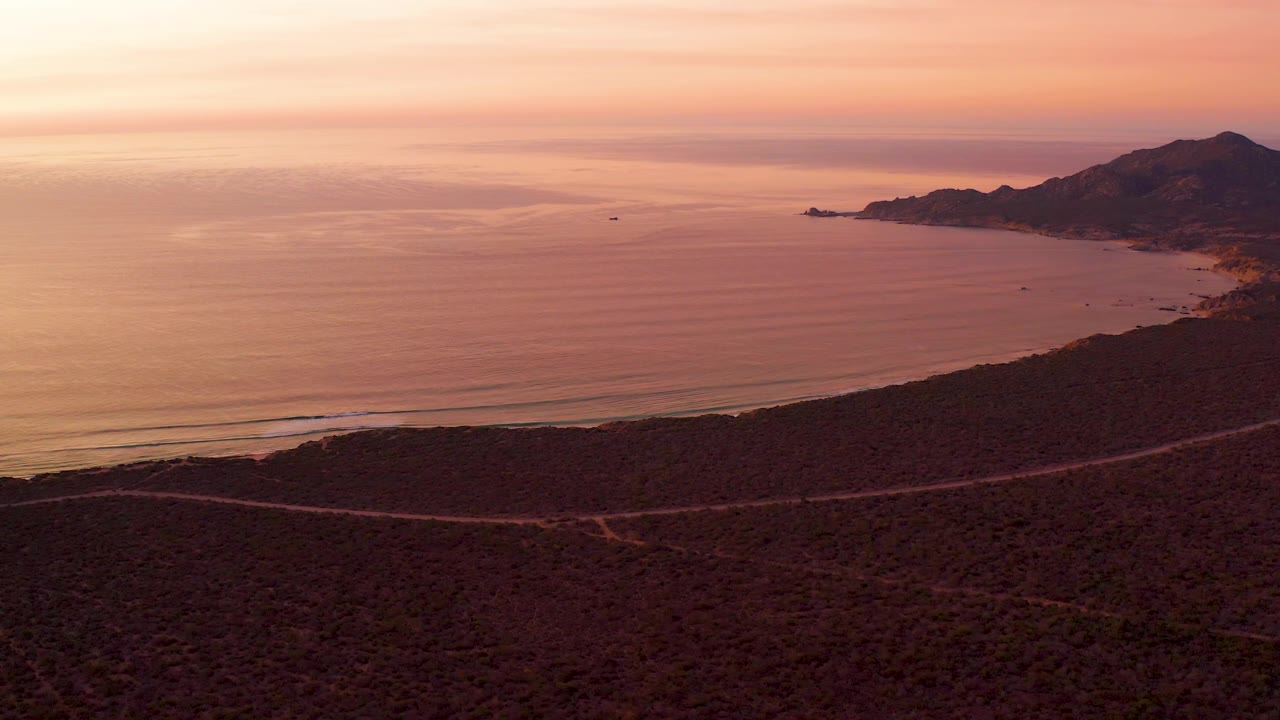 Aerial view in Cabo Pulmo, Baja California Sur with a pale sunset glow over the ocean, a curving shoreline below, a road cutting across the dark foreground hill, distant hills at right, and no visible people.