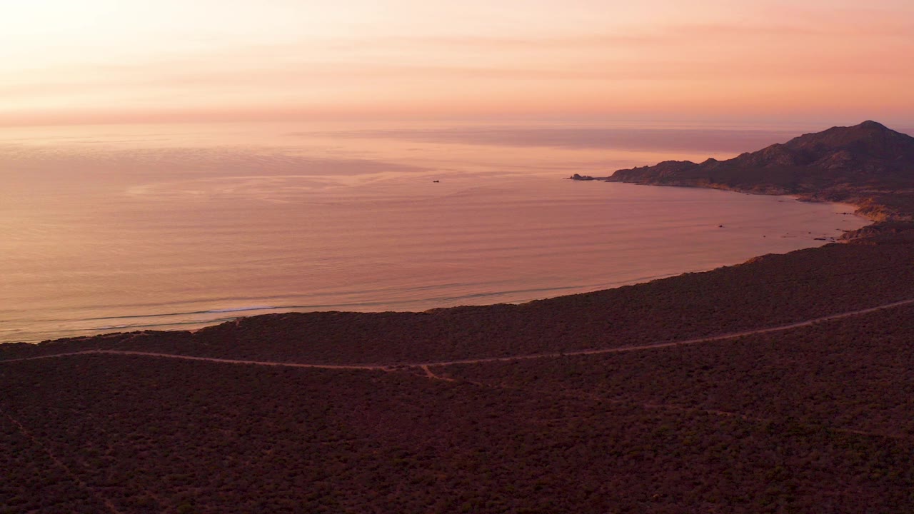 Wide aerial view of Cabo Pulmo at sunset with a scrub-covered hillside in front, a thin coastal road below, calm ocean water, distant mountains, and no visible people.