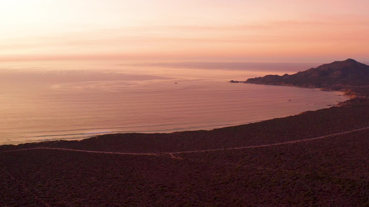 Wide sunset coastline with dry foreground, coastal road, ocean, and right-side mountains.