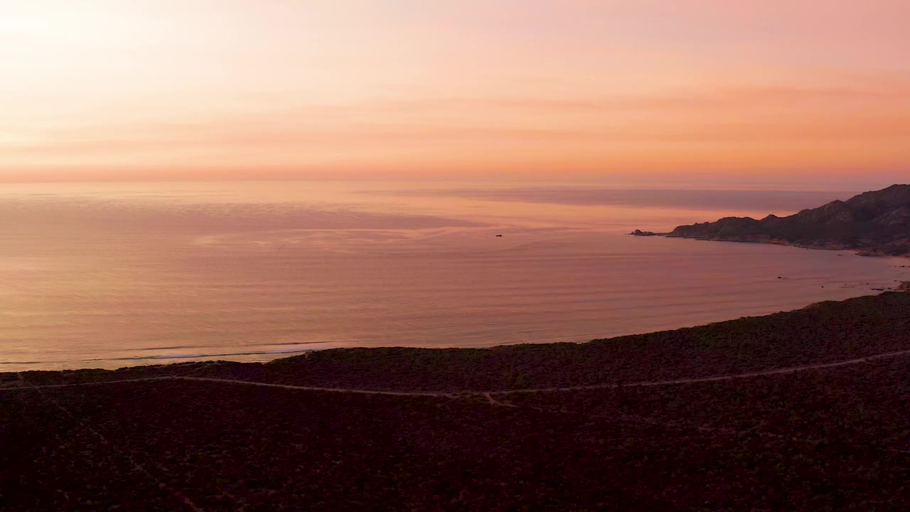 Aerial frame of the winding Cabo Pulmo shoreline road crossing the dark foreground while the ocean and pink sky fill most of the view, with no visible people.