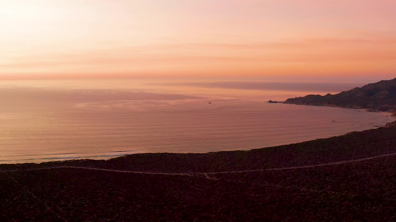A Cabo Pulmo sunset view shows the road running along the lower edge, ocean across the center, hills at right, and no visible people in the scene.