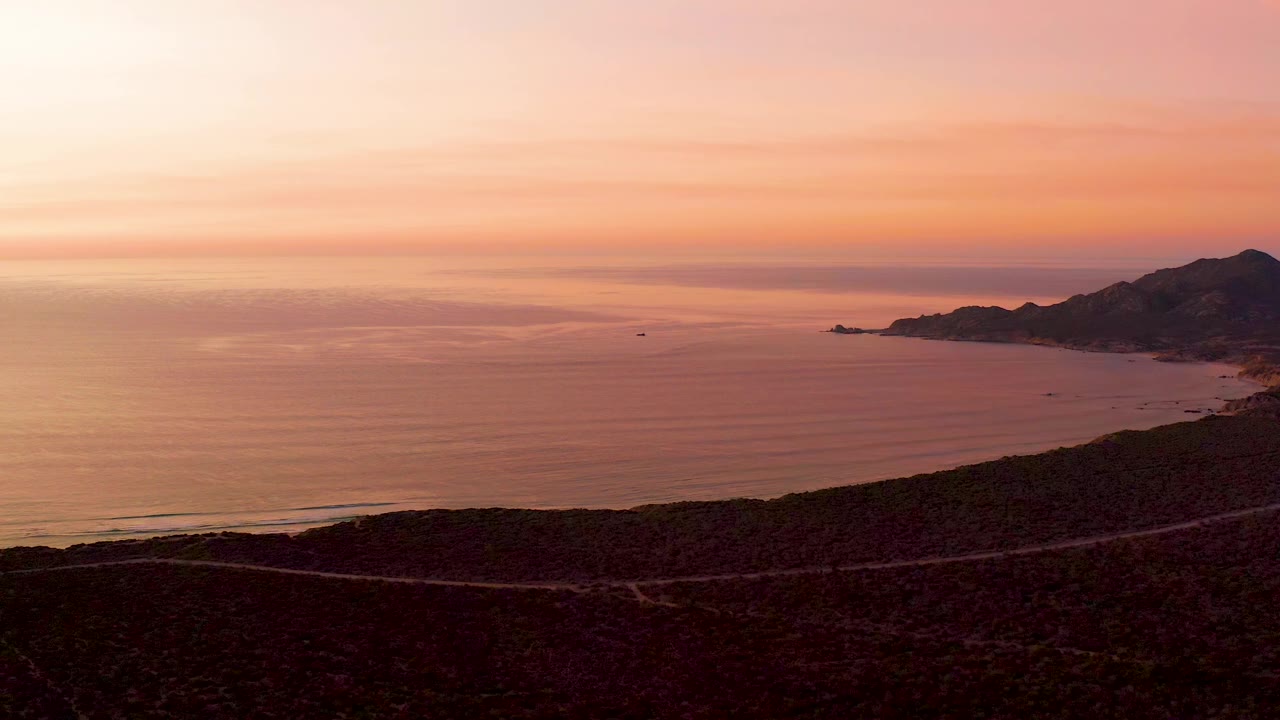 Cabo Pulmo sunset view with the mountain coastline on the right, a small boat on the open water, dark land in the foreground, and no visible people.