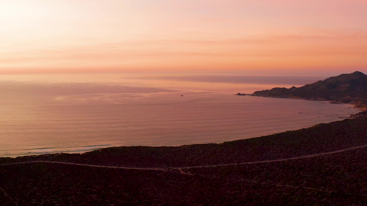 Cabo Pulmo aerial coastal view with a winding road across the dark foreground, smooth ocean water, distant boats, right-side hills, and no visible people.