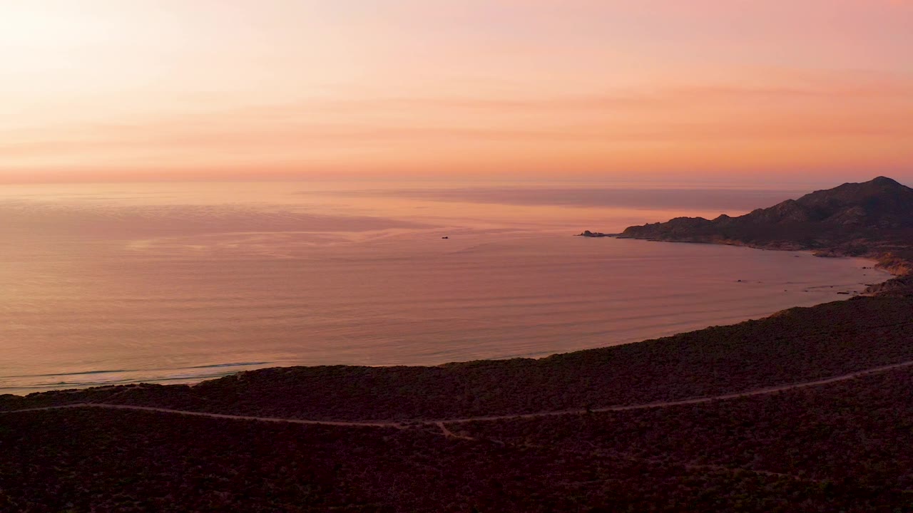 Cabo Pulmo sunset frame with the ocean filling most of the view, the shore road cutting through the dark lower foreground, distant hills to the right, and no visible people.