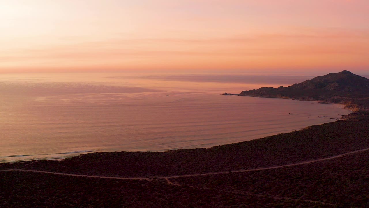 Cabo Pulmo coastal hillside at sunset with a winding road in the dark foreground, calm ocean water, distant hills to the right, and no visible people.