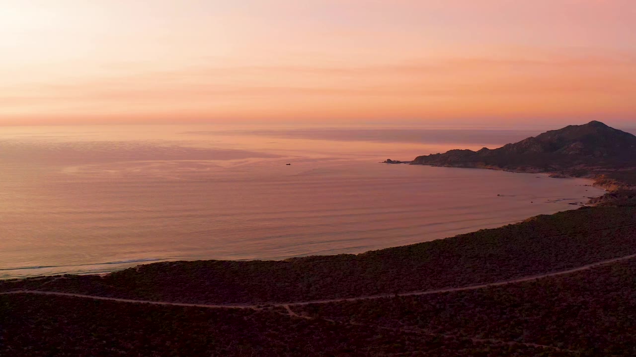 Cabo Pulmo sunset view with the hillside road crossing the lower frame, calm water filling the center, a mountain ridge on the right, and no visible people.
