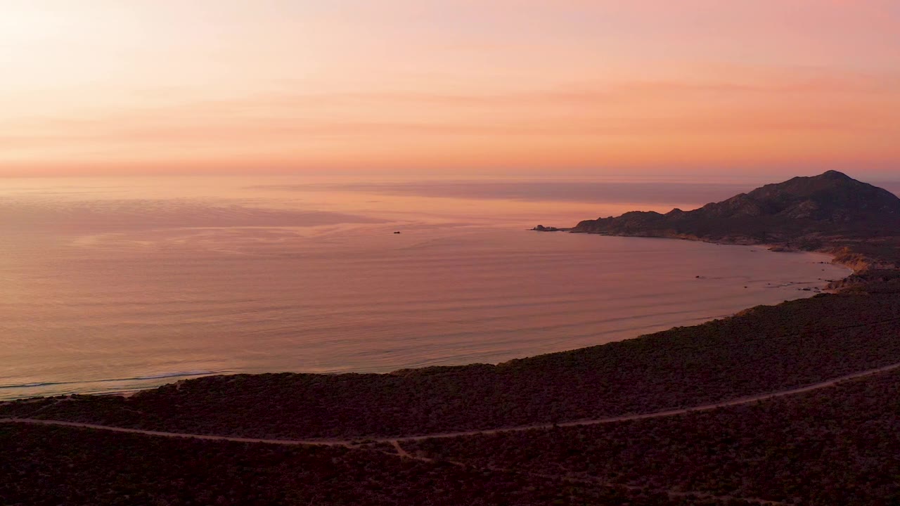 Ocean sunset with dark hillside foreground and distant mountain coast.