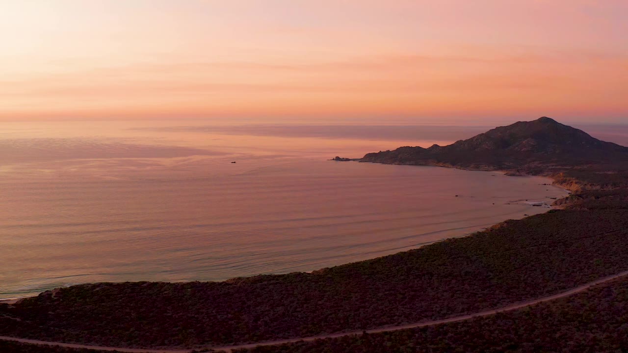 Elevated Cabo Pulmo sunset frame with broad calm water, a dark hillside foreground, a visible road near the bottom, mountain shoreline to the right, and no visible people.