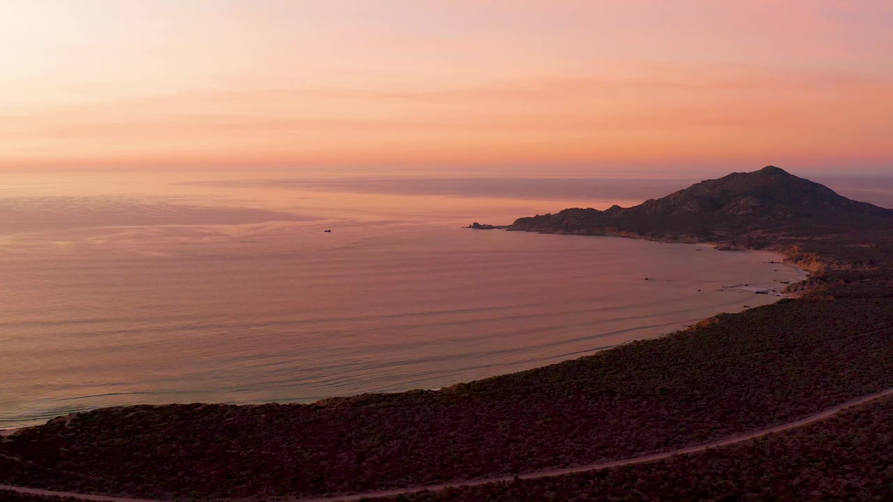 Cabo Pulmo coastline at dusk with still ocean water, a dark sloping foreground, a road tracing the hillside, mountain landform on the right, and no visible people.
