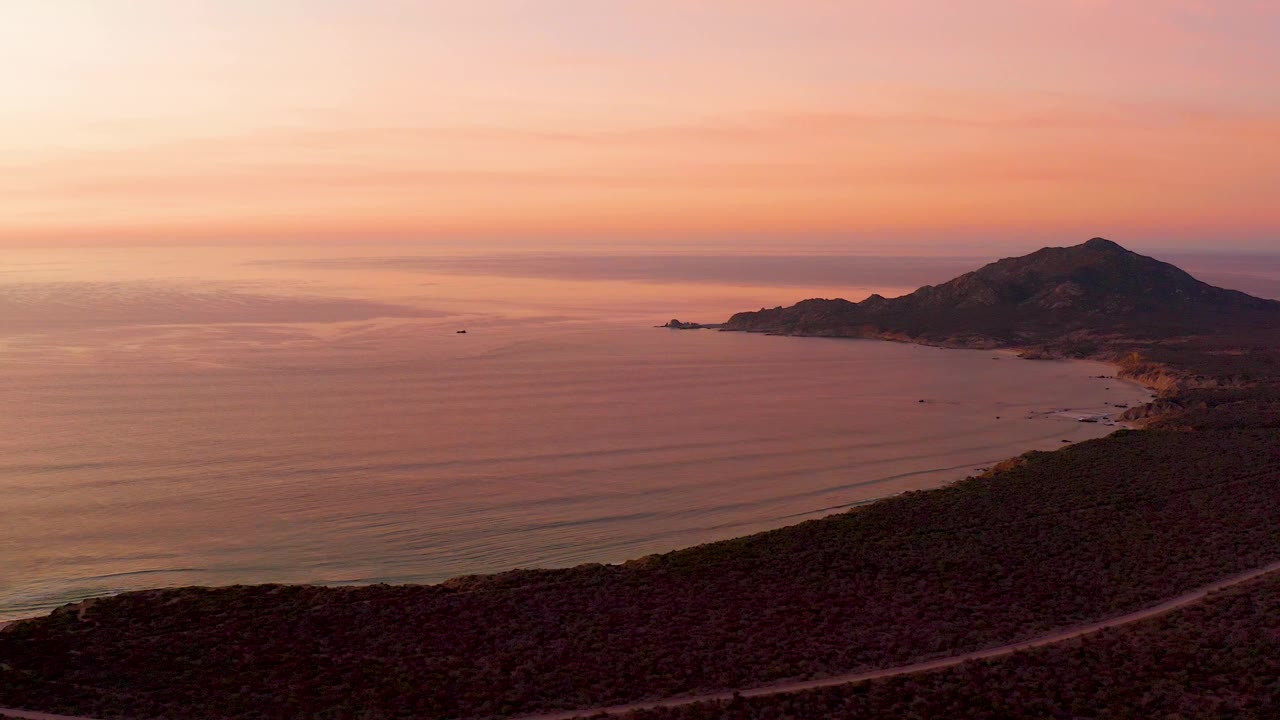Sunset coast with calm water, mountain, and road across hillside.