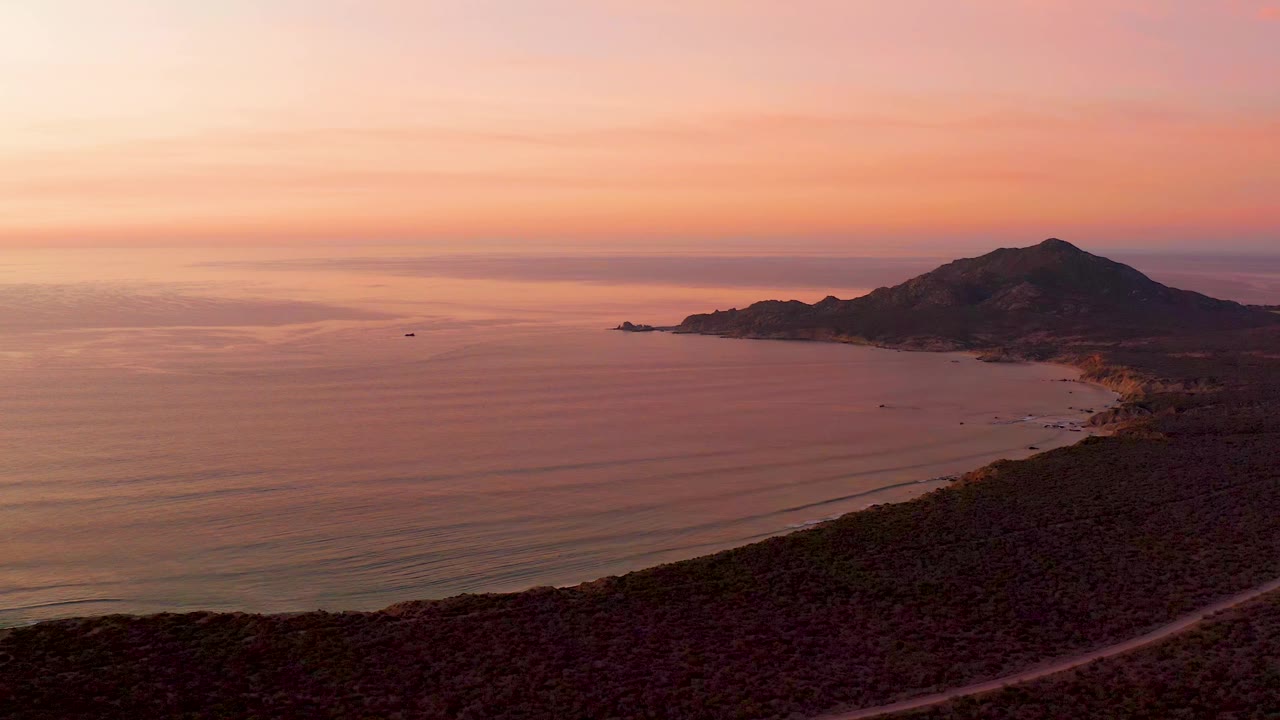 Cabo Pulmo coast at 0:01 with the horizon held high, pink evening bands over calm water, the mountain pushed to the right, a foreground hillside road, a curved beach, a small boat offshore, and no visible people.