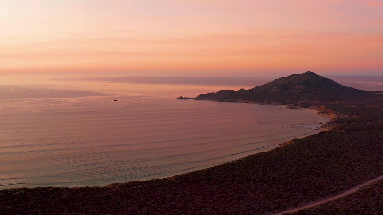 Cabo Pulmo coastline at 0:03 with the mountain silhouetted against a pink sky, calm water in the bay, the hillside road dark across the foreground, beach detail along the shore, a small boat offshore, and no visible people.