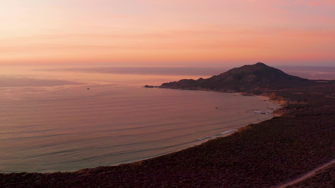 Aerial view over Cabo Pulmo at sunset, with a small boat offshore, low ocean ripples, a curving beach, dark scrubland in the foreground, and no visible people.