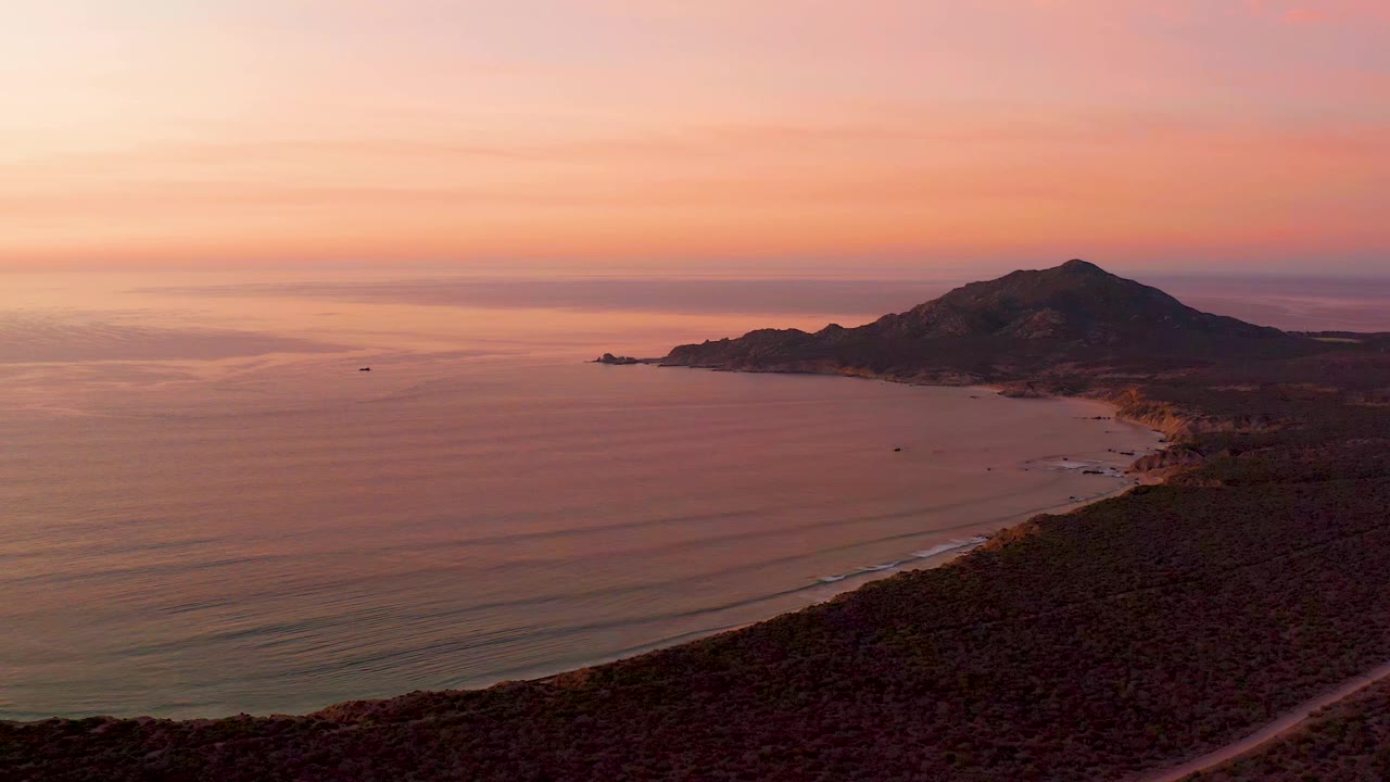 Cabo Pulmo coastline seen from above at 0:01, with the small boat left of center, smooth sunset water, a beach tracing the shore, and no visible people.