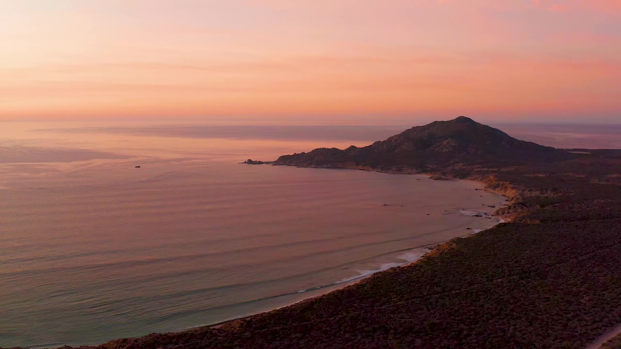 Cabo Pulmo shoreline seen from above at sunset, with the ocean spreading left, the beach line running toward a dark hill, soft cloud bands, and no visible people.