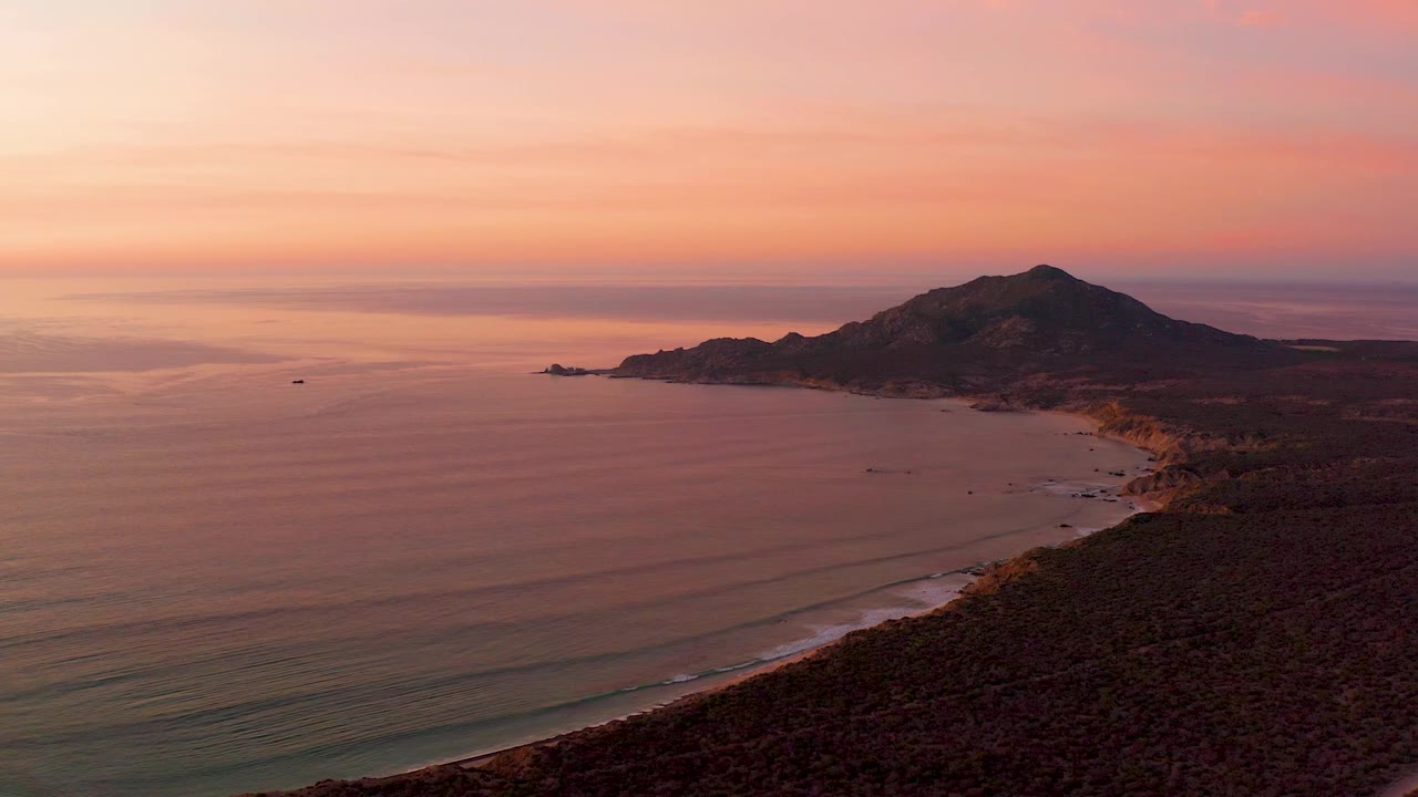 Wide Cabo Pulmo coastal frame with the water taking most of the left side, a curved sandy shore at the base of the hills, layered sunset clouds, and no visible people.