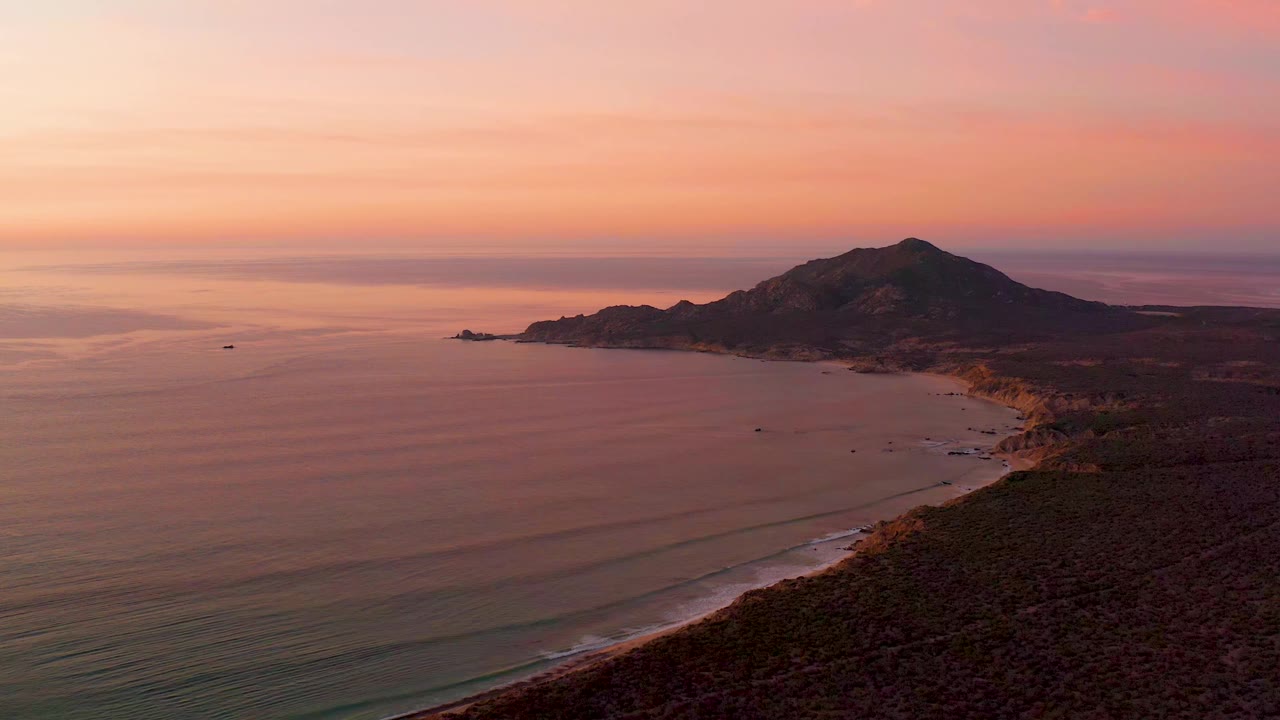 Cabo Pulmo coastline seen from above with parallel wave lines approaching the sandy beach, dark land on the right, a hill in the distance, and no visible people.