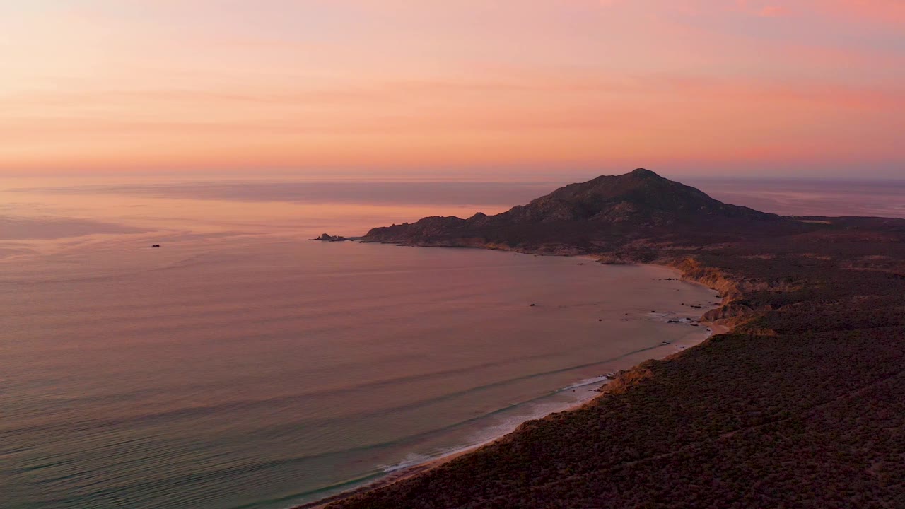 A high aerial frame over Cabo Pulmo shows smooth ocean water on the left, a narrow sandy shore on the right, mountain terrain beyond, and no visible people.