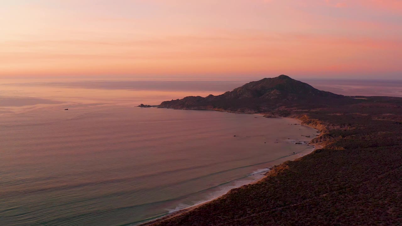 Cabo Pulmo shoreline fills the lower right while low wave bands cross the water, the hill sits near the horizon, and the sunset sky is pink and orange with no visible people.