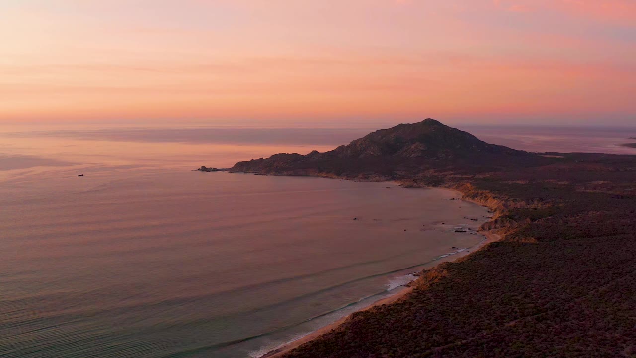 Cabo Pulmo coastline at sunset with smooth ocean water, faint wave bands near shore, a sandy beach along the right side, and no visible people.