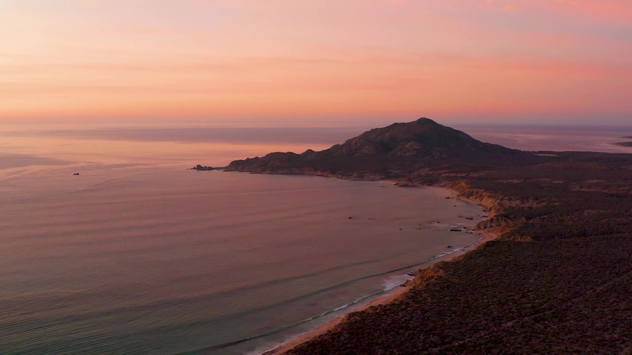 Aerial Cabo Pulmo sunset frame with the ocean filling the left foreground, the beach curving into the right midground, the hill set back on the coast, and no visible people.