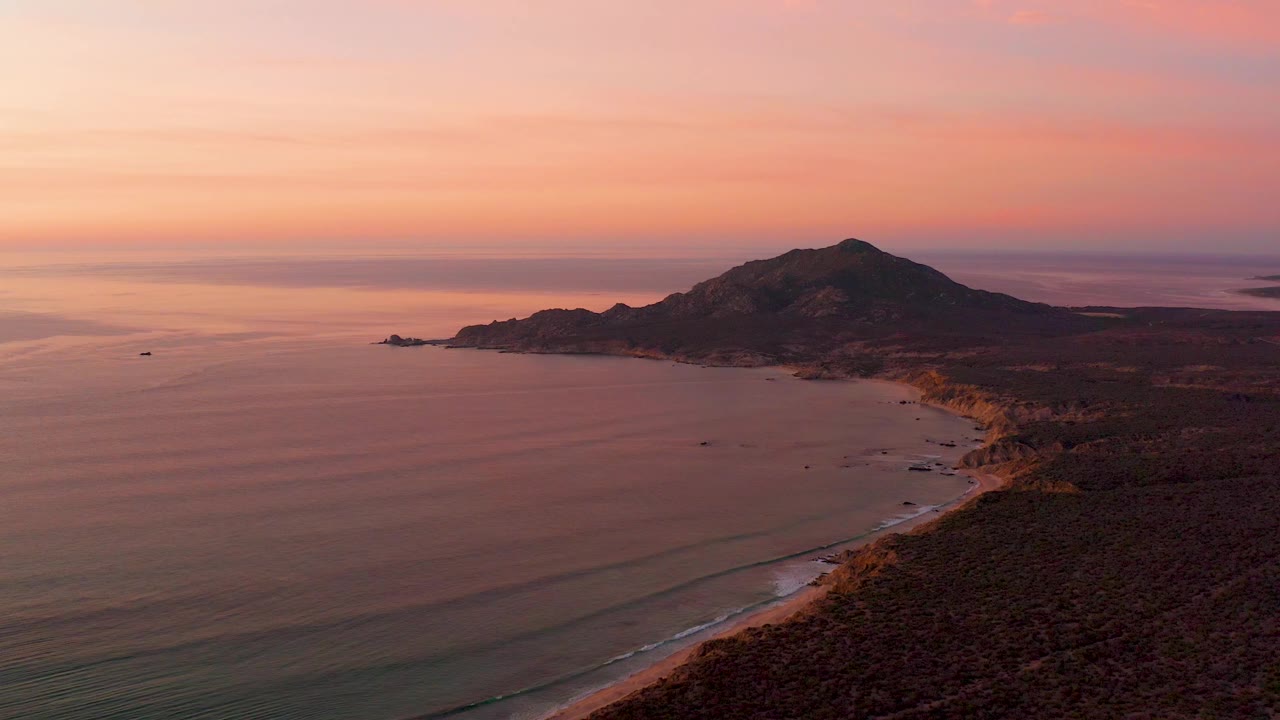 Cabo Pulmo aerial sunset view with low parallel waves approaching the beach, dark land on the right, the hill beyond the shore, and no visible people.