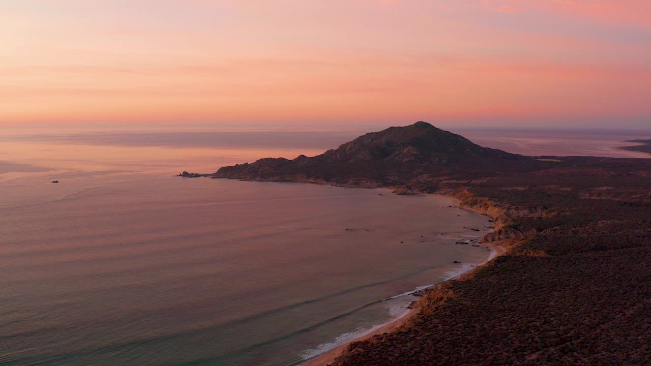 Aerial view over Cabo Pulmo at sunset, with calm ocean on the left, a rocky shoreline curving toward dark hills, pink-orange sky, and no visible people.