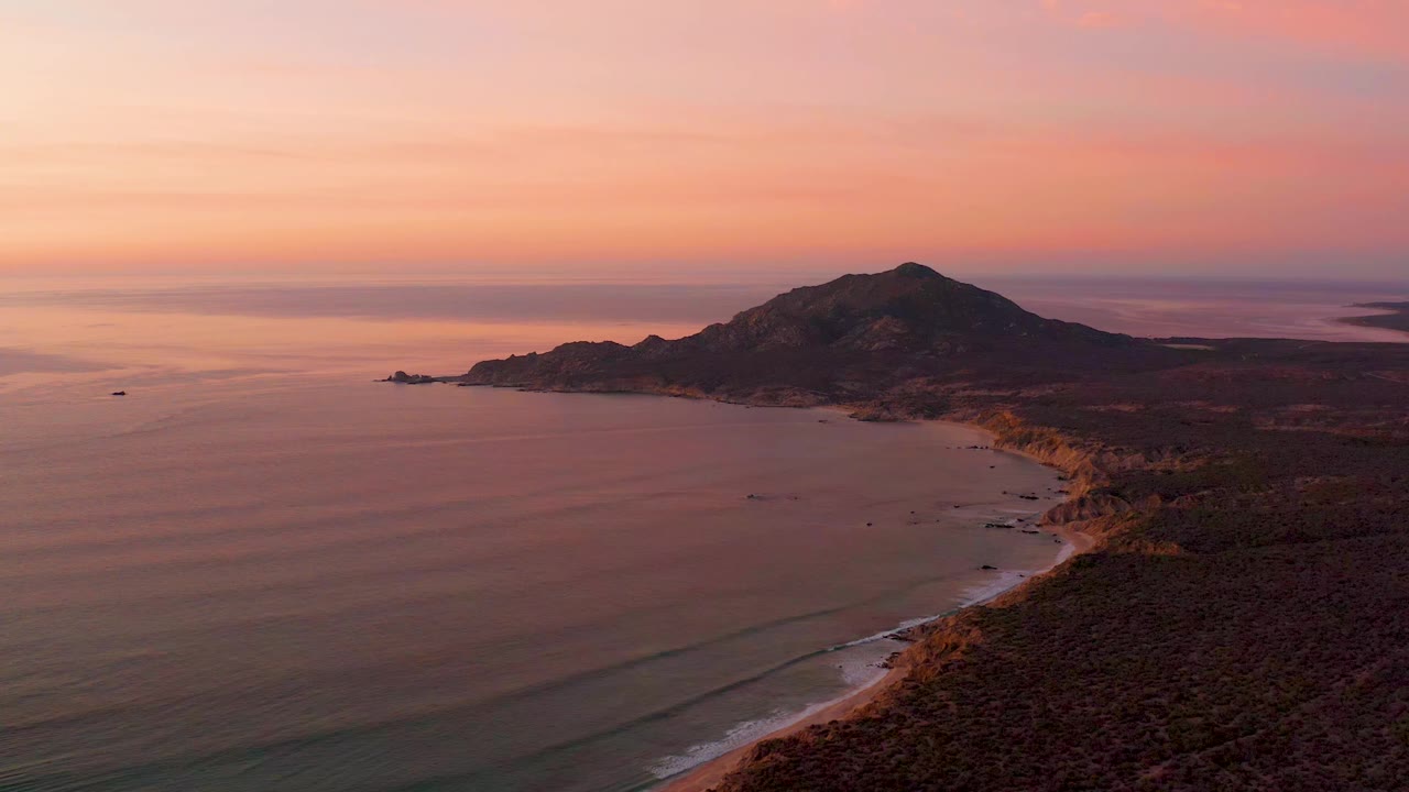 Cabo Pulmo coastline seen from above, with soft orange light across the sky, faint wave bands on the water, rugged shore at right, distant hills, and no visible people.