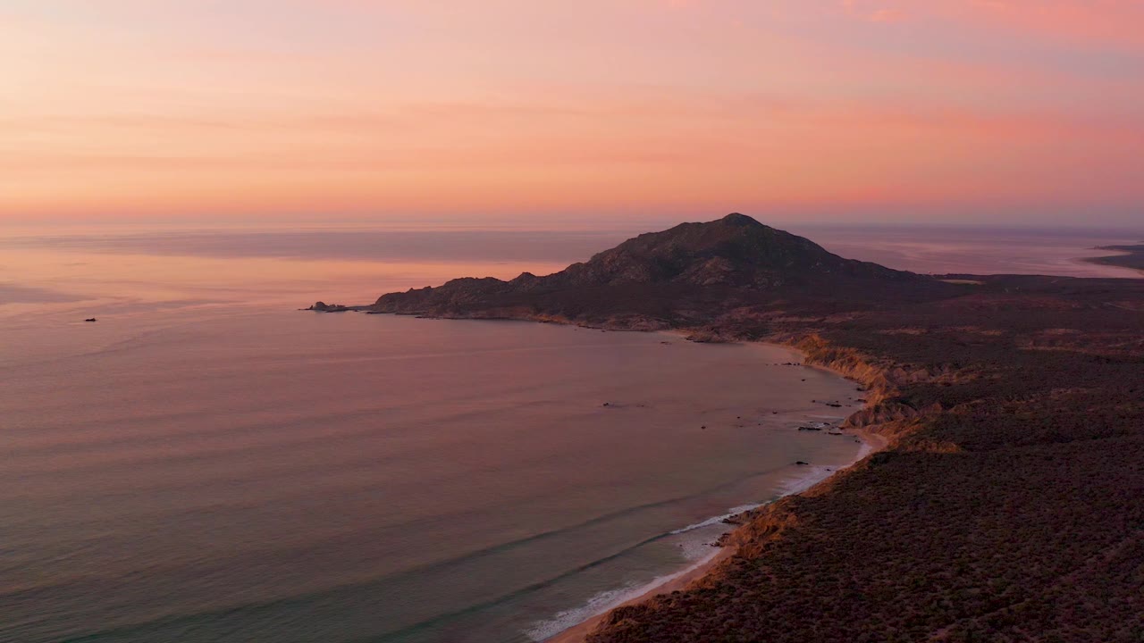 Wide aerial frame of Cabo Pulmo with calm ocean filling the left side, low waves approaching the beach, a rocky headland and hills in the background, and no visible people.